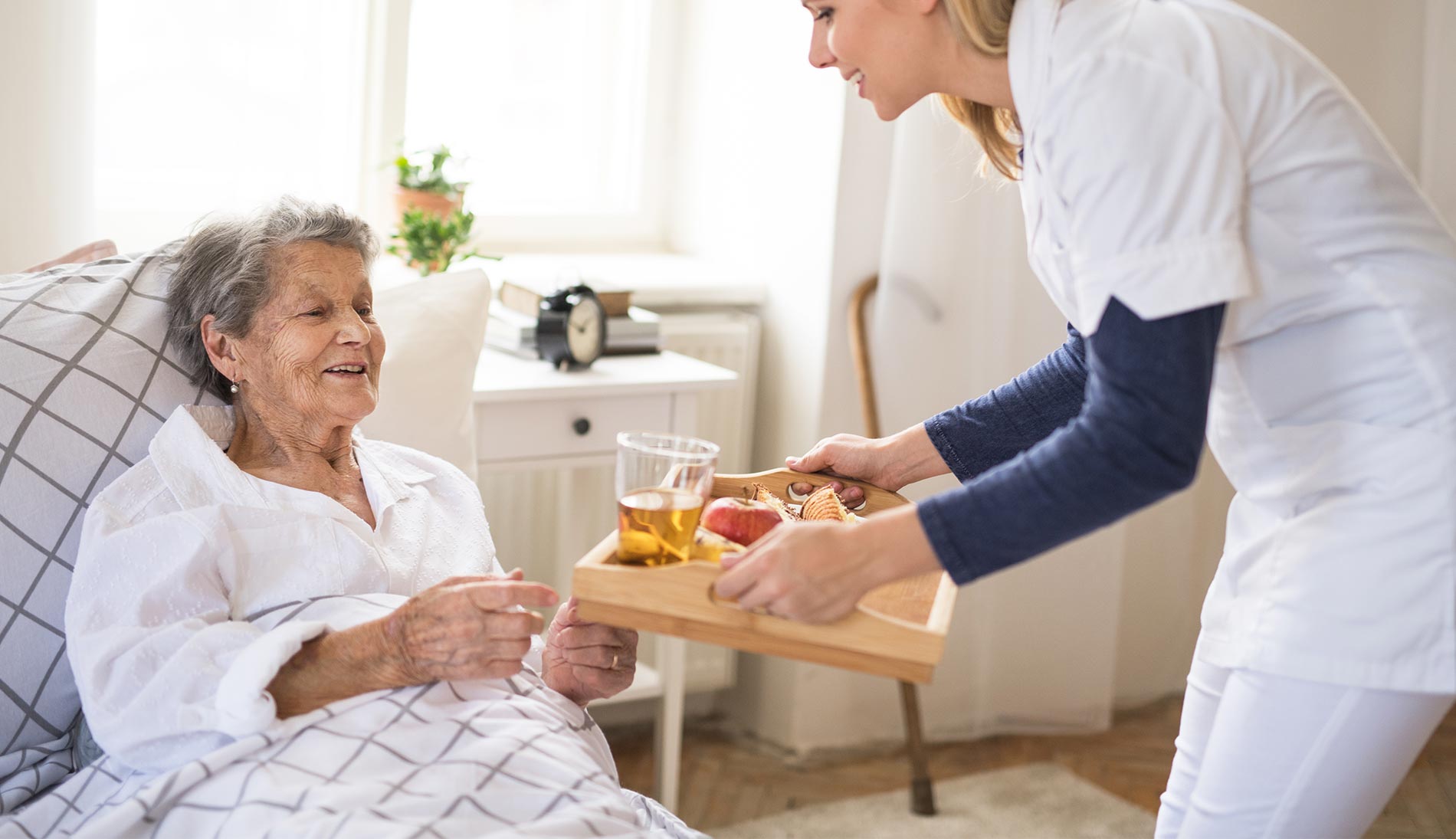 Pflegekraft bringt einer Seniorin das Frühstück ans Bett und unterstützt sie bei der morgendlichen Versorgung.