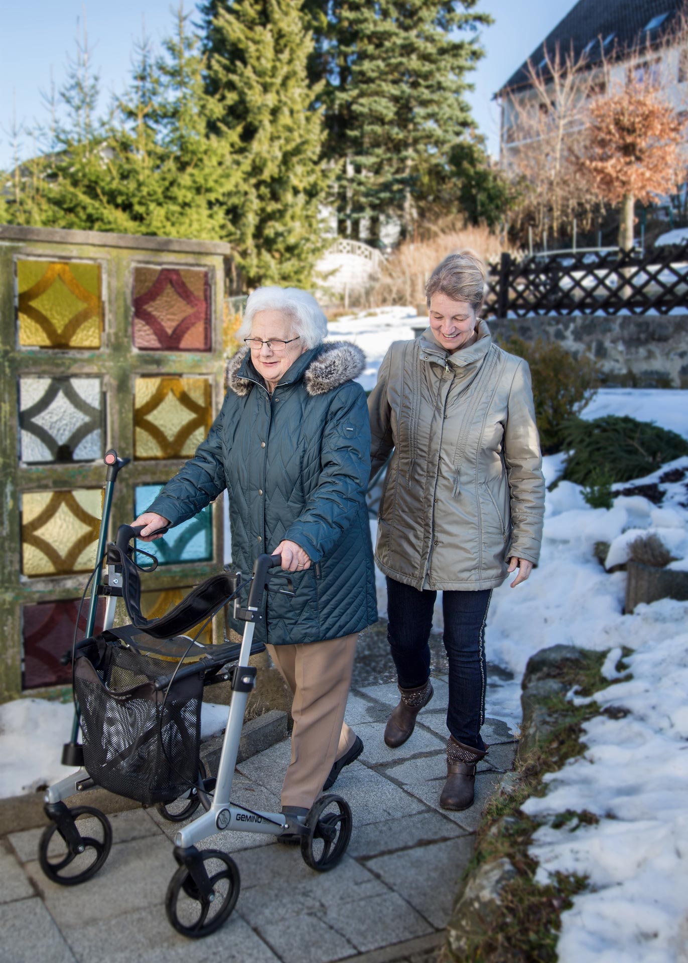 Pflegekraft begleitet eine Seniorin mit Rollator bei einem Spaziergang im Freien und unterstützt ihre Mobilität im Alltag.