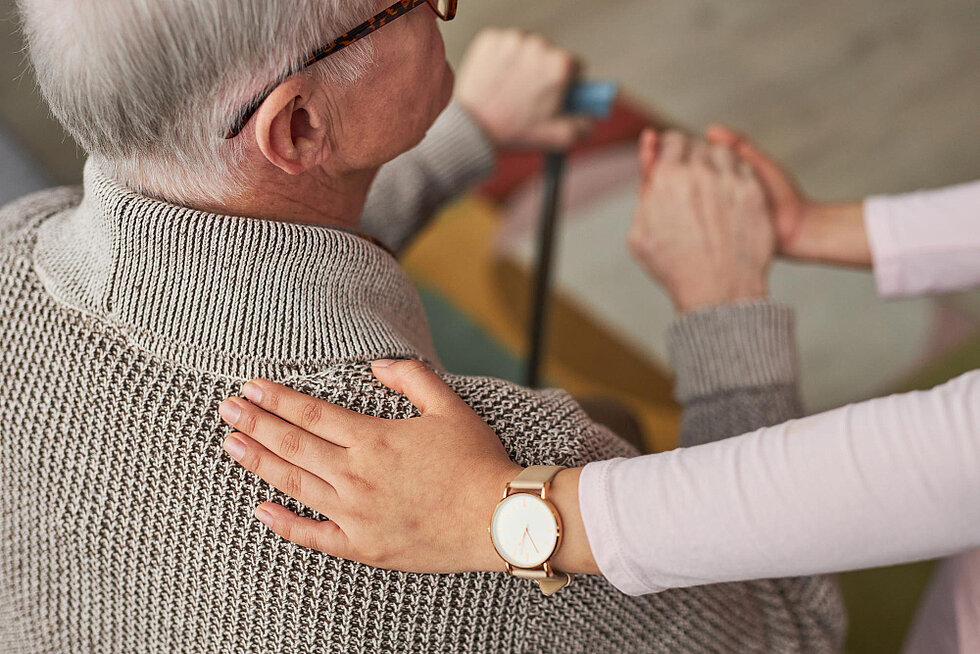 Pflegekraft legt einem Senior unterstützend die Hand auf die Schulter und begleitet ihn im Alltag.