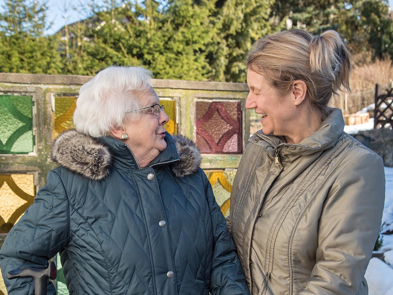 Pflegekraft und Seniorin lächeln sich bei einem gemeinsamen Spaziergang an und unterhalten sich miteinander.