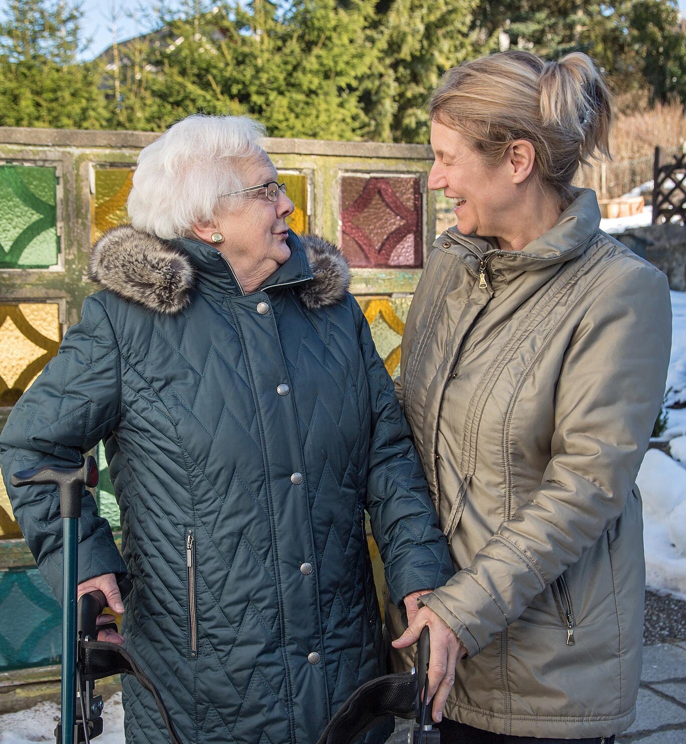 Pflegekraft und Seniorin lächeln sich bei einem gemeinsamen Spaziergang an und unterhalten sich miteinander.