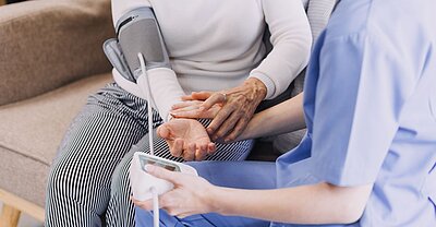 Pflegekraft misst bei einem Hausbesuch den Blutdruck einer älteren Person und kontrolliert die Vitalwerte.