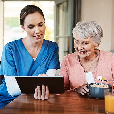 Pflegekraft zeigt einer Seniorin Inhalte auf einem Tablet und unterstützt sie bei der Beratung im häuslichen Umfeld.