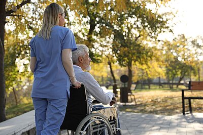 Pflegekraft begleitet einen Senior im Rollstuhl bei einem Spaziergang im Park.