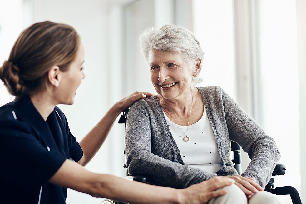 Pflegekraft spricht mit einer Seniorin im Rollstuhl und begleitet sie in einem persönlichen Gespräch.